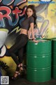 A woman leaning on a green barrel in front of a graffiti covered wall.